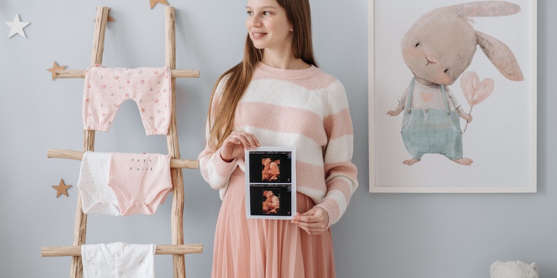 Tea collection Vs Hanna Andersson-Expecting mum standing in nursery holding a scan from the baby while next to baby clothes hanging out on a ladder.
